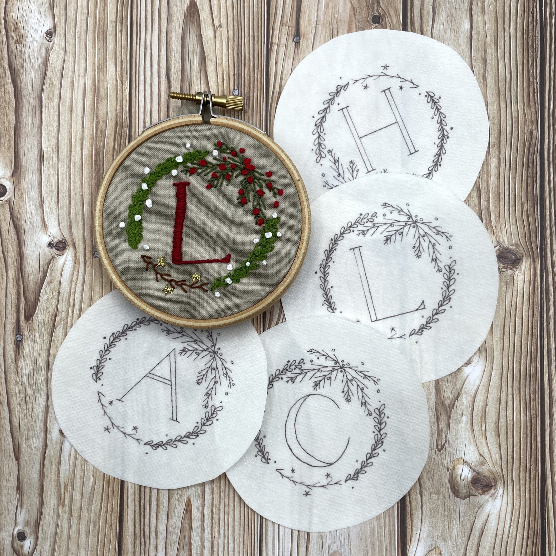 Pack of Christmas wreath embroidery stickers and finished embroidered version: a mini wreath with greenery, branches and red and white berries. Initial "L" stitched in the centre.