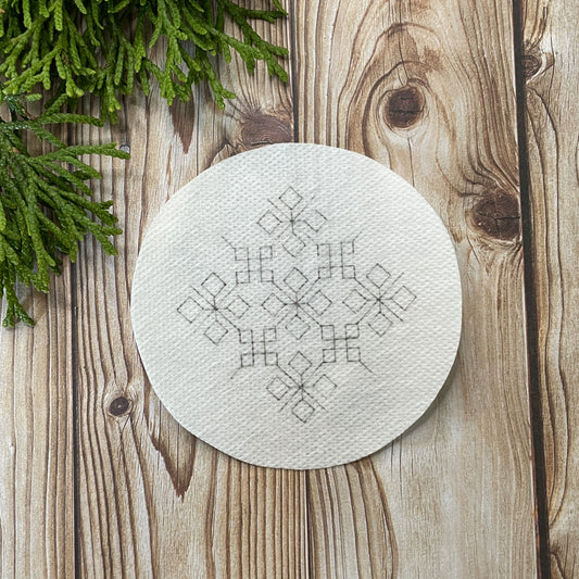 Circular embroidery sticker with a geometric snowflake design on a wooden surface with Christmas greenery.